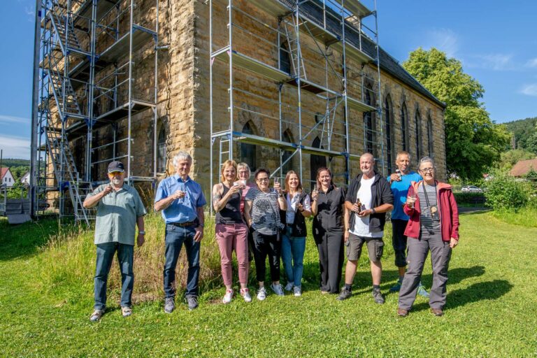 Mit einem Glas Sekt wird die erfolgreiche Sanierung gemeinsam gefeiert – Vertreter:innen der Gemeinde, Beteiligte und Unterstützer:innen vereint vor der Kirche Merkers.